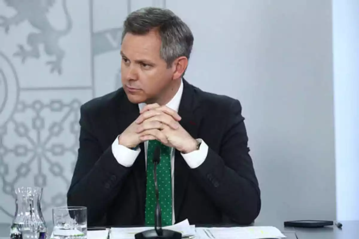 Hombre de traje oscuro y corbata verde con puntos blancos, sentado en una mesa con documentos, un micrófono, un vaso de agua y una jarra, con las manos entrelazadas y una expresión seria.