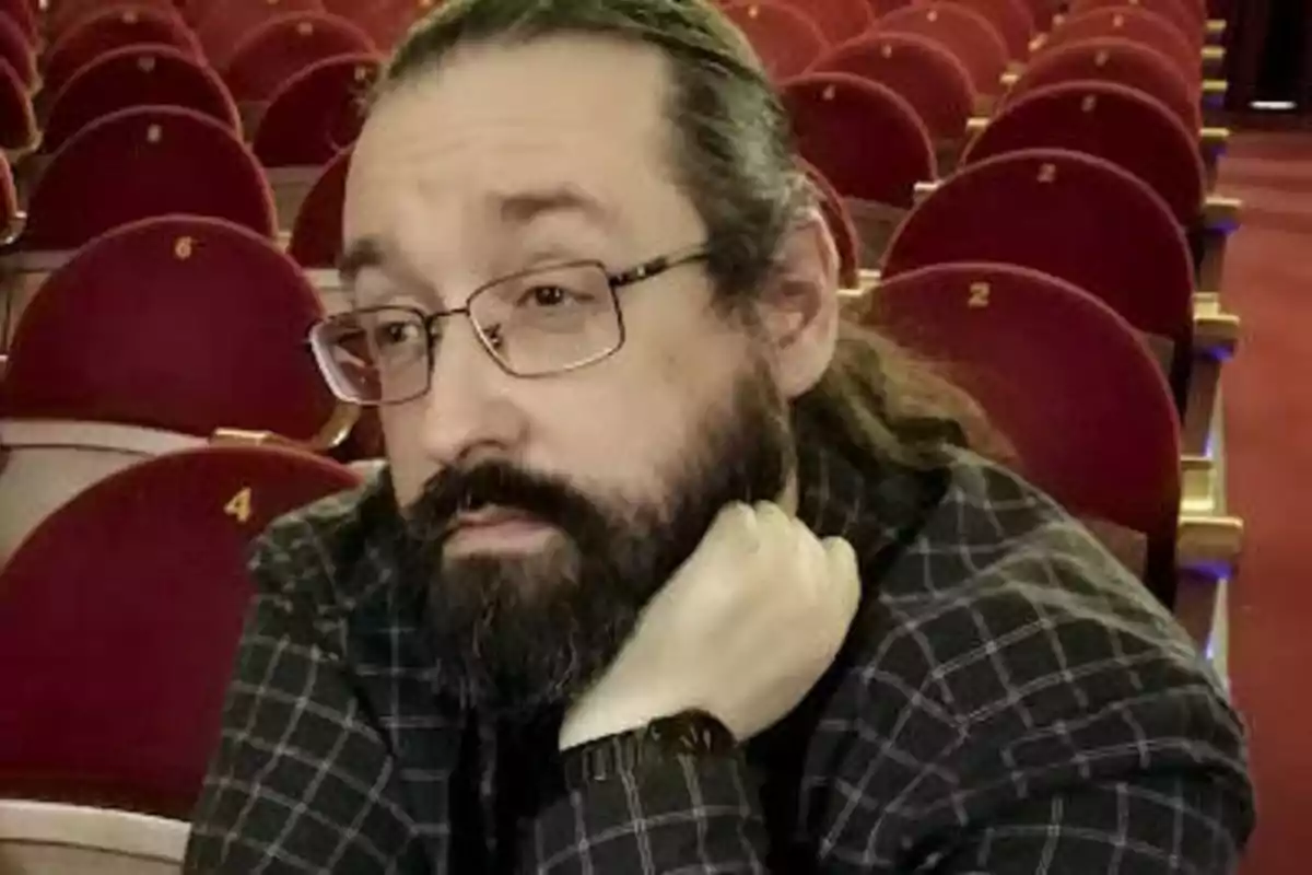 Un hombre con barba y gafas, sentado en una sala de teatro con asientos rojos numerados.