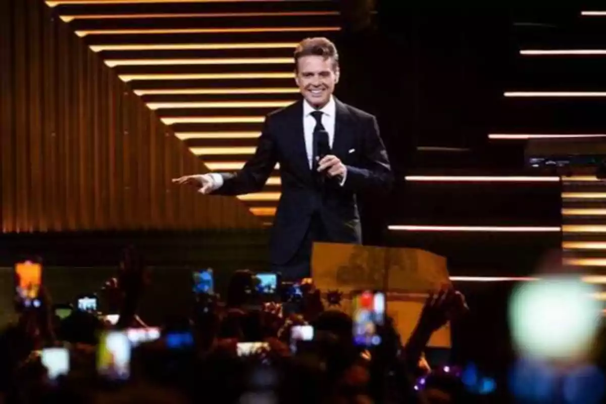 Un hombre con traje negro y corbata canta en un escenario iluminado mientras el público lo graba con sus teléfonos.