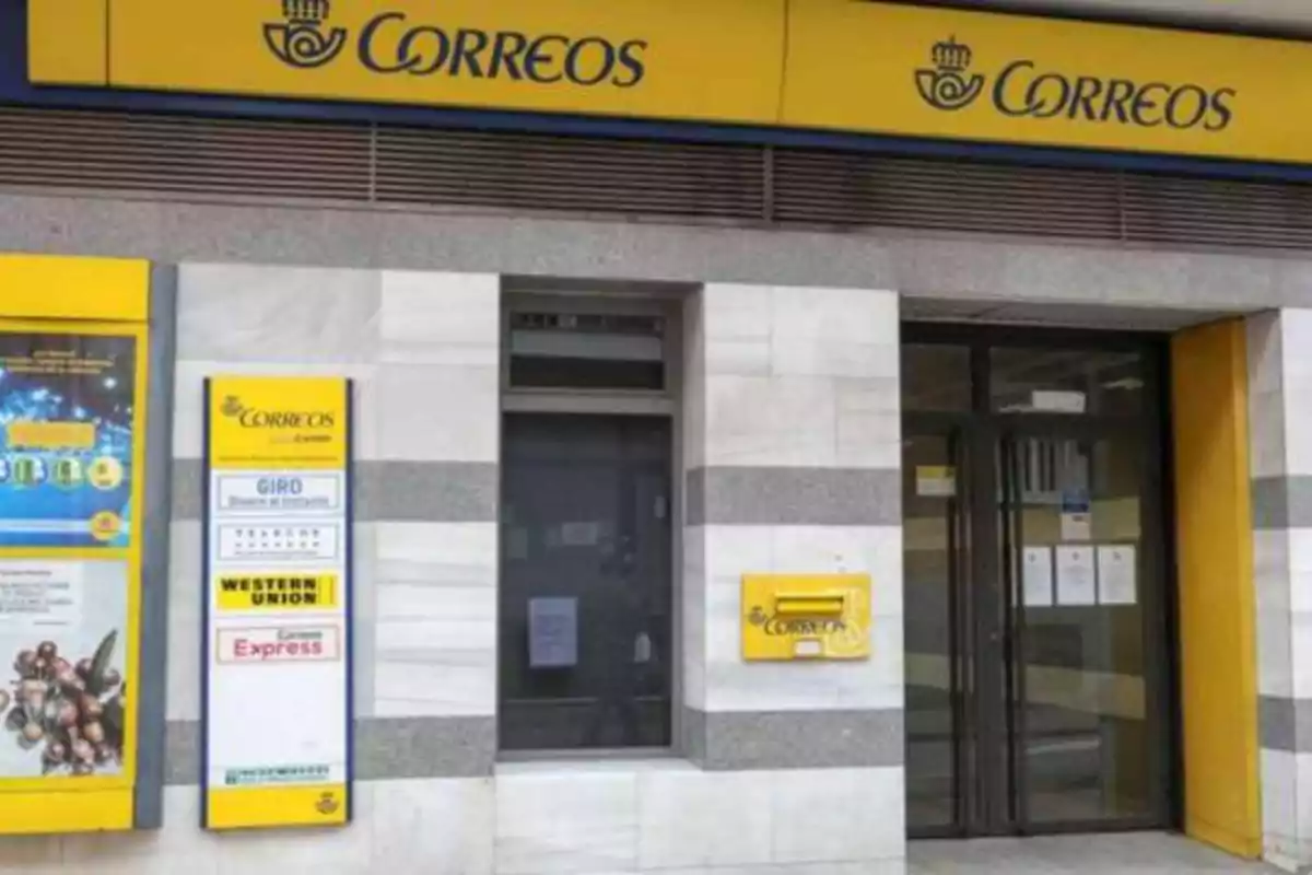 La imagen muestra la fachada de una oficina de Correos en España, con un letrero amarillo y azul en la parte superior que dice "Correos" y varios carteles informativos en la pared, incluyendo servicios de Western Union y Express.