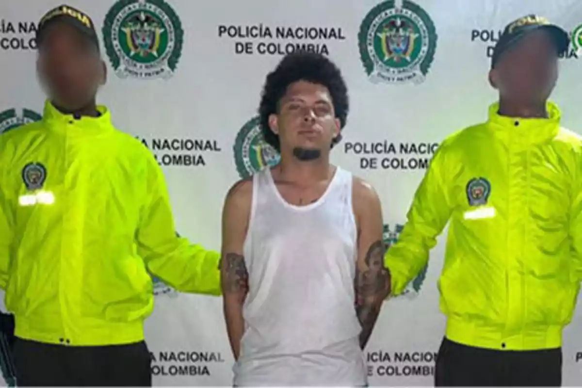 Un hombre con camiseta blanca es escoltado por dos oficiales de la Policía Nacional de Colombia que llevan chaquetas amarillas.