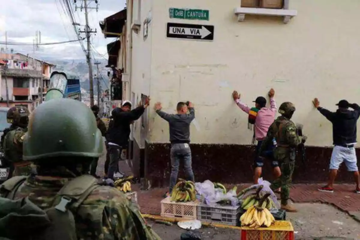 Soldados detienen a varias personas contra una pared en una calle urbana, con cajas de plátanos en el suelo.