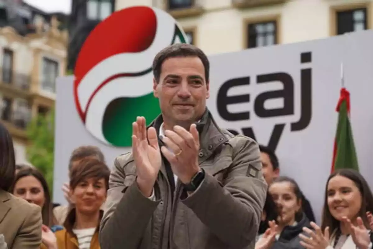 Un hombre aplaudiendo en un evento público con un grupo de personas detrás y un cartel con el logo de EAJ-PNV.