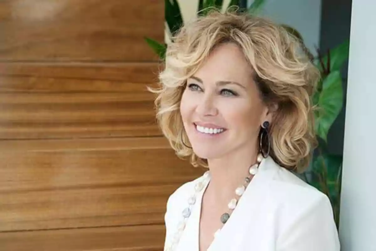 Mujer rubia sonriendo con un collar de perlas y fondo de madera.