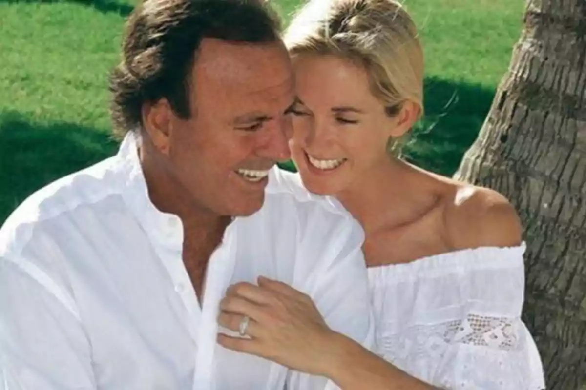 Una pareja sonriente vestida de blanco se abraza al aire libre junto a un árbol.