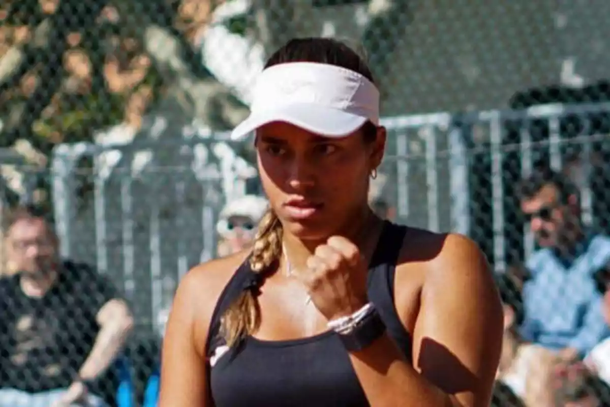 Una tenista con una visera blanca y una camiseta sin mangas negra celebra con el puño cerrado en una cancha de tenis al aire libre.