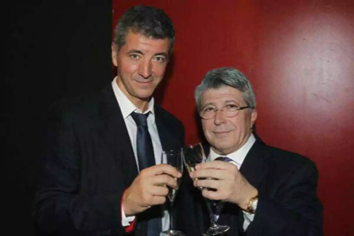 Dos hombres vestidos de traje brindando con copas de champán frente a un fondo rojo.