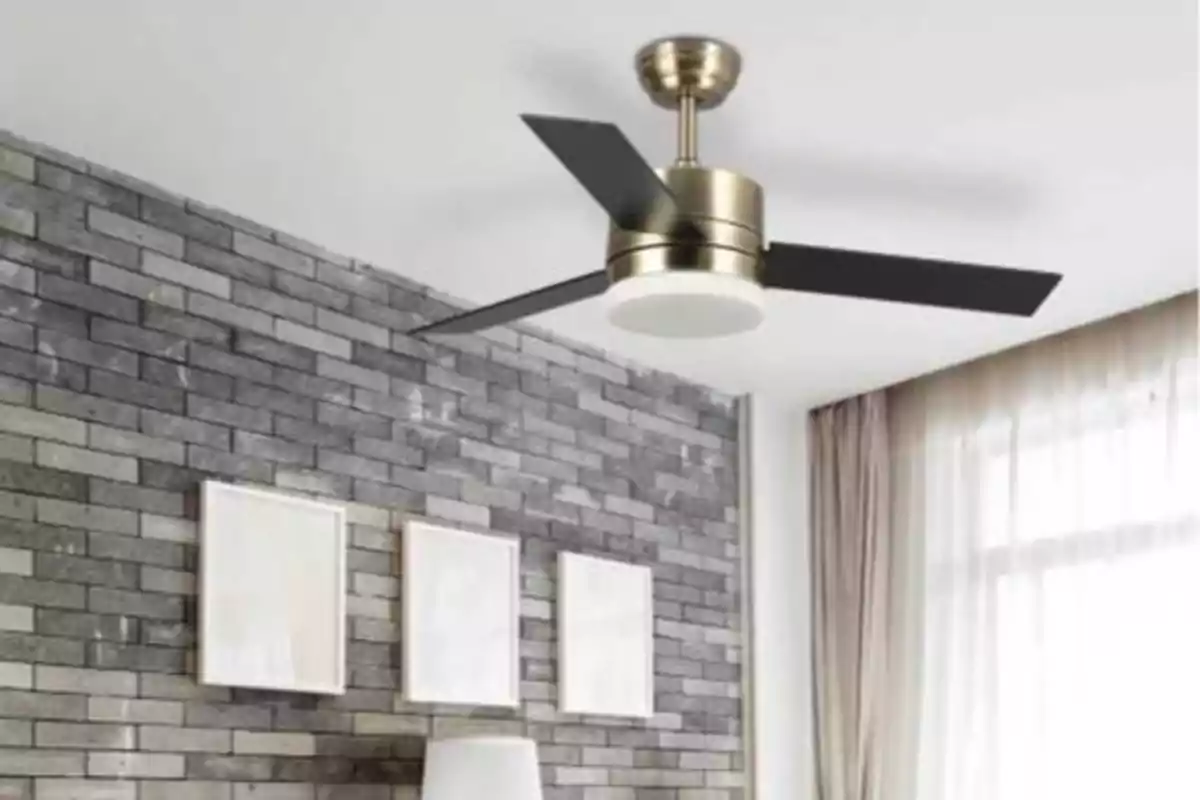 Ventilador de techo moderno con tres aspas negras en una habitación con pared de ladrillo gris, tres cuadros enmarcados y una lámpara de pie blanca.