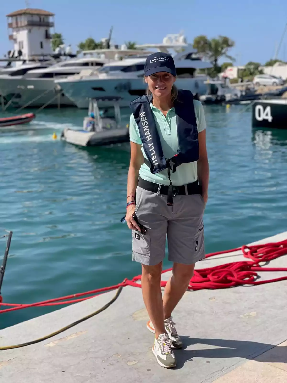 Una mujer con chaleco salvavidas y gorra está de pie en un muelle con varios yates y botes en el fondo.