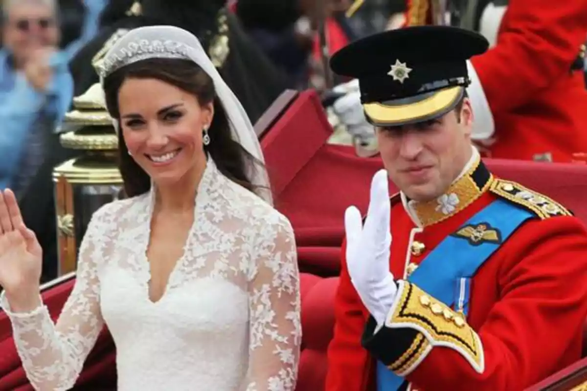 Una pareja vestida de manera formal, con la mujer usando un vestido de novia blanco y el hombre un uniforme militar rojo, ambos saludando y sonriendo.