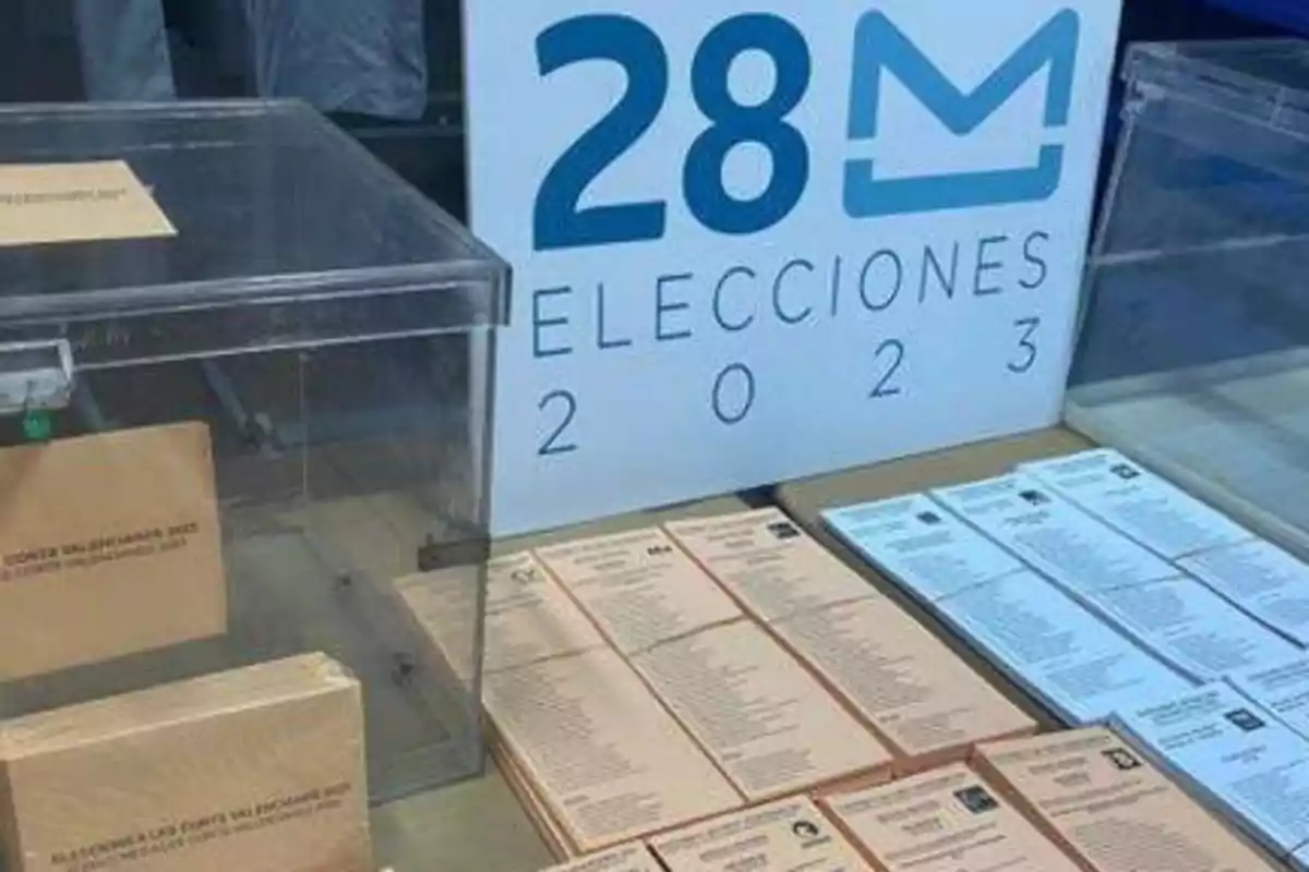 Urnas y papeletas de votación junto a un cartel que dice "28M Elecciones 2023".