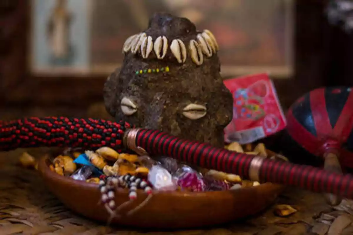Una figura de piedra decorada con conchas y cuentas, rodeada de diversos objetos rituales en un plato.
