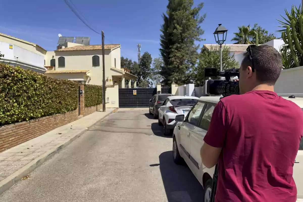 Un camarógrafo con camiseta roja graba en una calle residencial con coches estacionados y una casa de fondo. Un camarógrafo con camiseta roja graba en una calle residencial con coches estacionados y una casa de fondo.