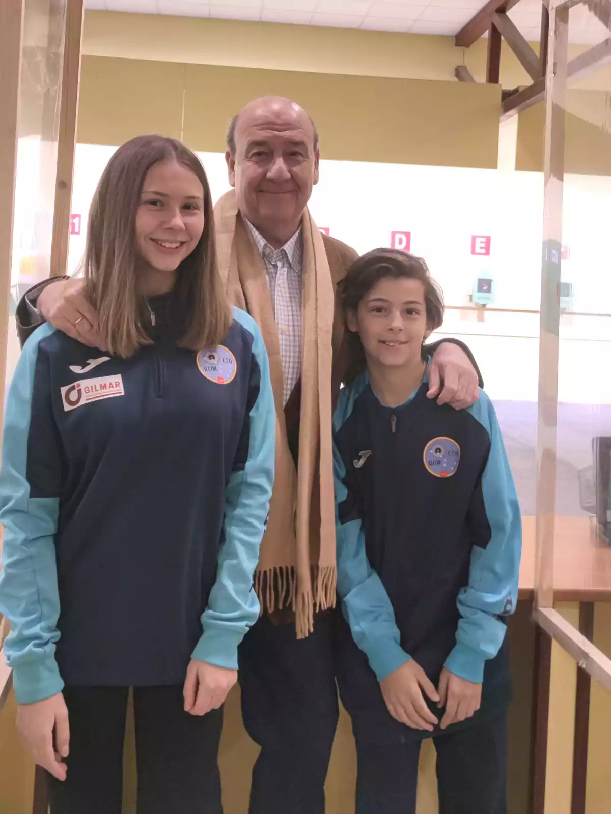 Tres personas posando juntas, dos jóvenes con uniformes deportivos y un adulto con bufanda, en un entorno interior.