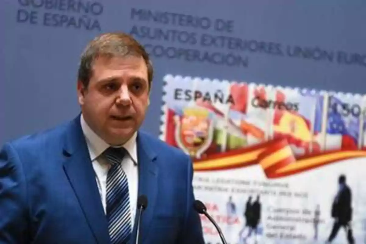 Un hombre con traje azul y corbata a rayas habla frente a un micrófono, con un fondo que muestra un cartel del Gobierno de España y el Ministerio de Asuntos Exteriores, Unión Europea y Cooperación.