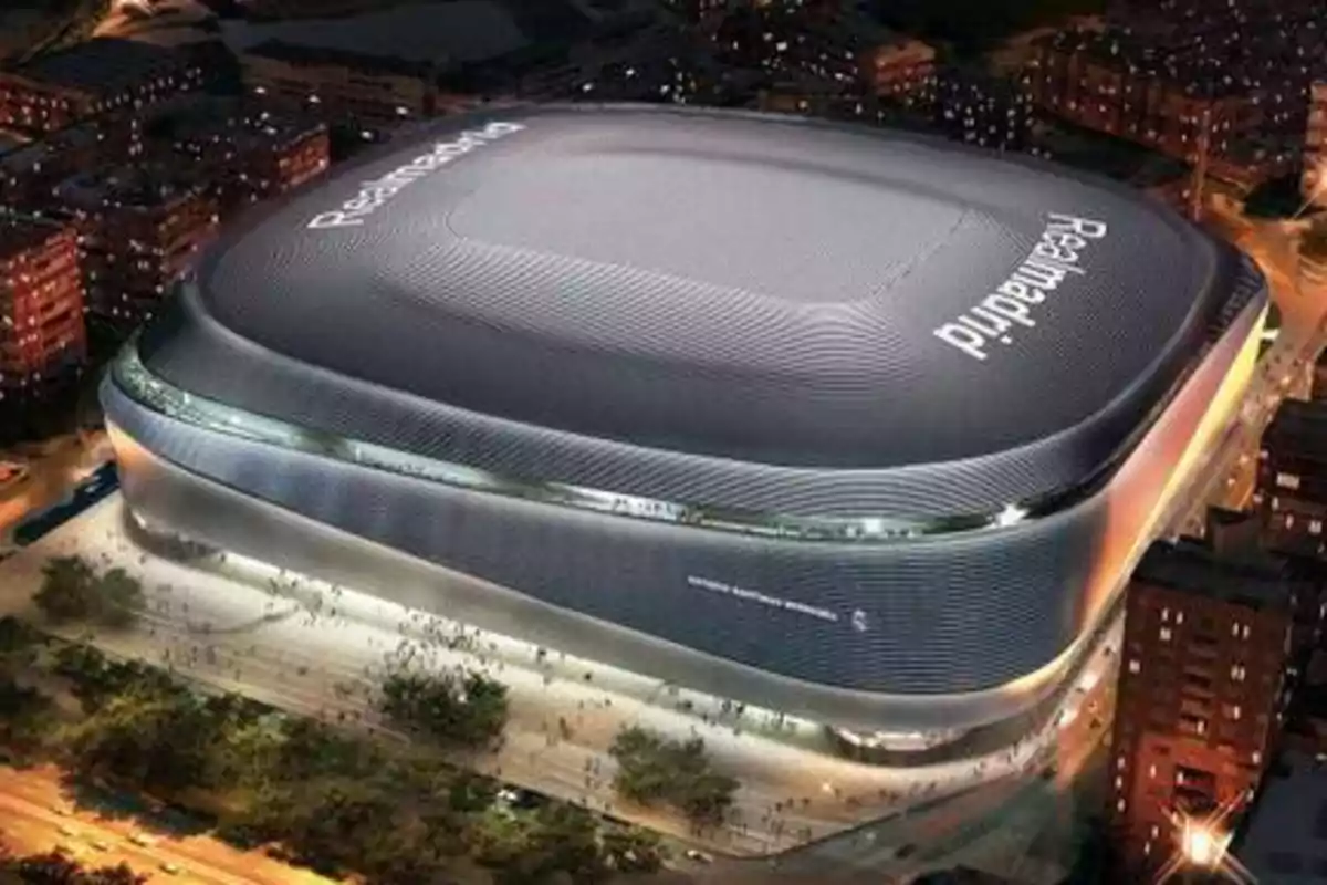 Vista aérea de un estadio moderno iluminado por la noche con el nombre "Real Madrid" visible en el techo.