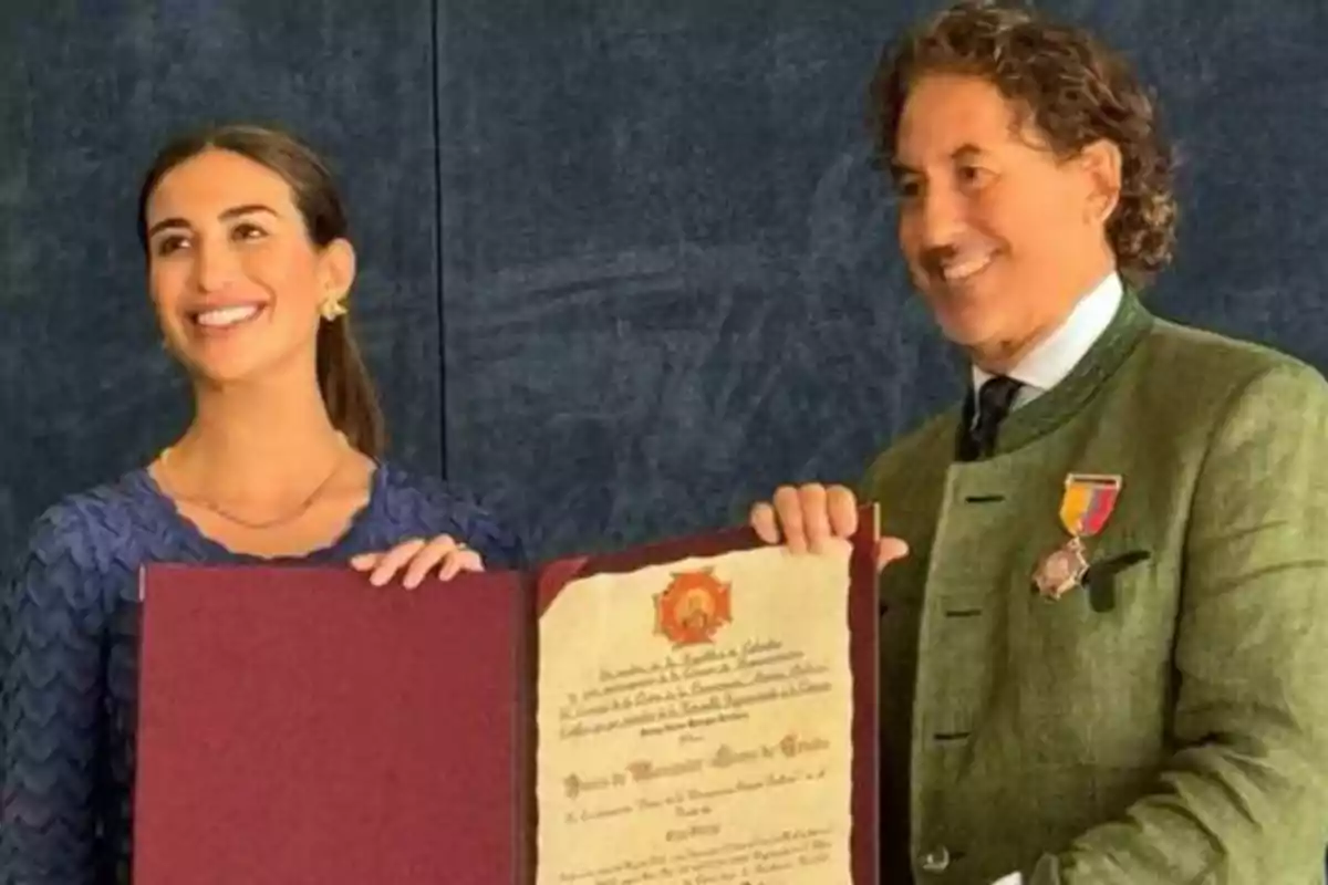 Dos personas sonrientes sosteniendo un diploma o certificado enmarcado, una mujer a la izquierda con una blusa azul y un hombre a la derecha con un traje verde y una medalla en la solapa.