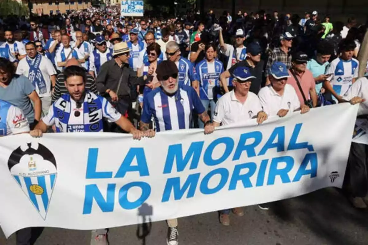 Un grupo de personas con camisetas y bufandas del equipo de fútbol CD Alcoyano marchan sosteniendo una pancarta que dice "LA MORAL NO MORIRÁ".
