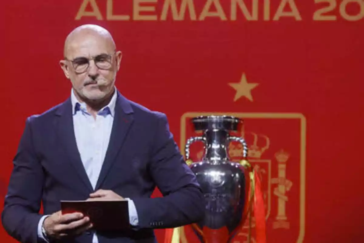 Un hombre con gafas y barba, vestido con un traje oscuro y camisa blanca, sostiene un documento frente a un fondo rojo con la palabra "ALEMANIA" y un trofeo plateado detrás de él.