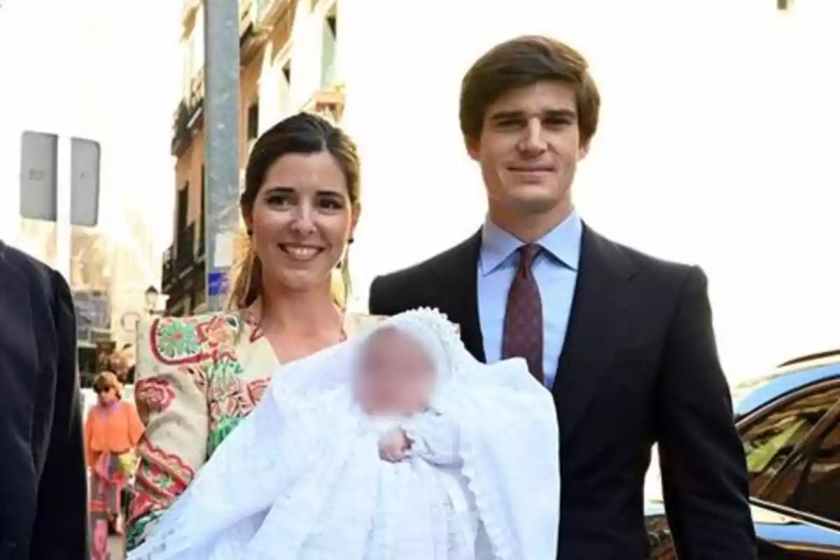 Una pareja sonriente sostiene a un bebé vestido de blanco en una calle urbana.