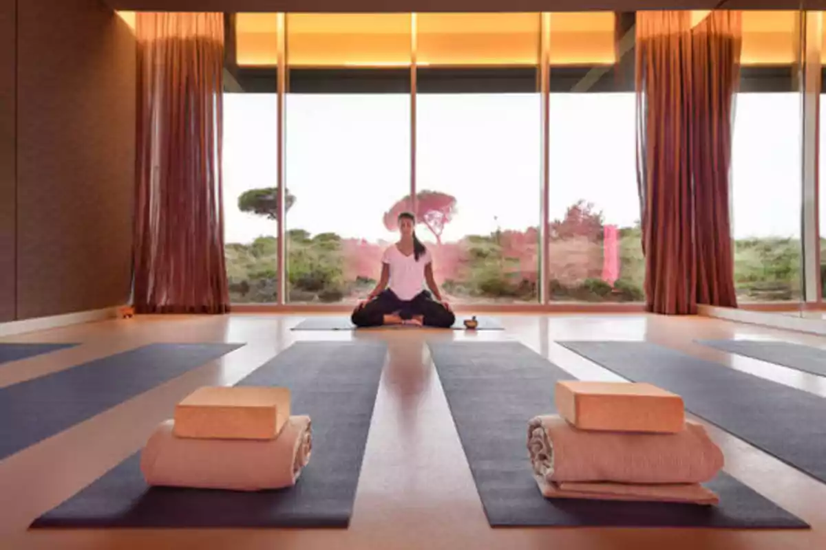 Persona practicando yoga en una sala con grandes ventanales y colchonetas en el suelo.