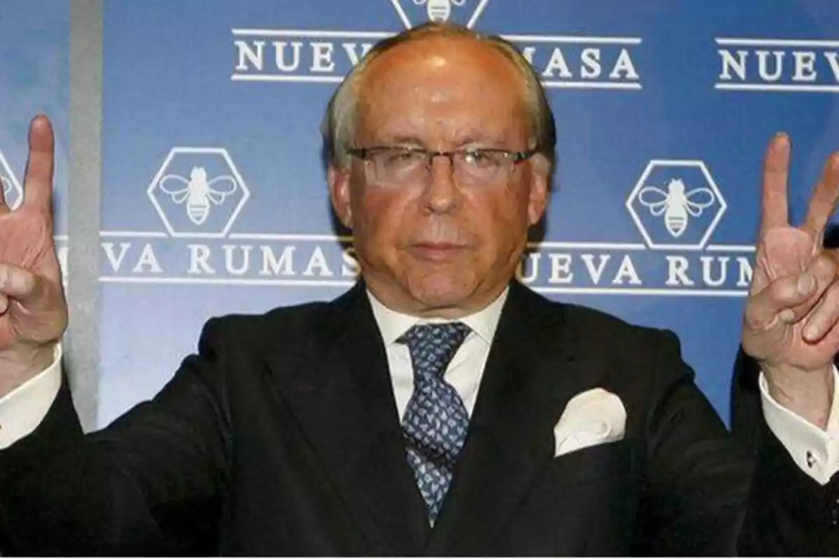 Hombre con traje y corbata haciendo el signo de la paz con ambas manos frente a un fondo con el logo y texto "Nueva Rumasa".