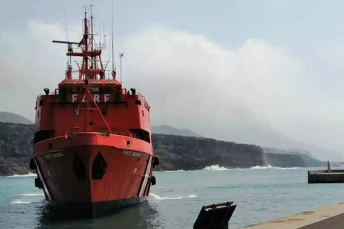 Un barco rojo se aproxima al muelle con un paisaje montañoso y nublado al fondo.