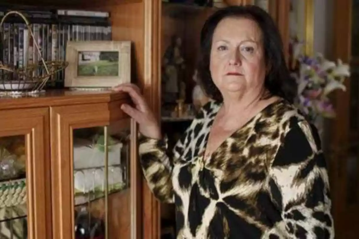 Una mujer de pie junto a un mueble de madera con puertas de vidrio, sosteniendo la esquina del mueble con una mano, vestida con una blusa de estampado animal.