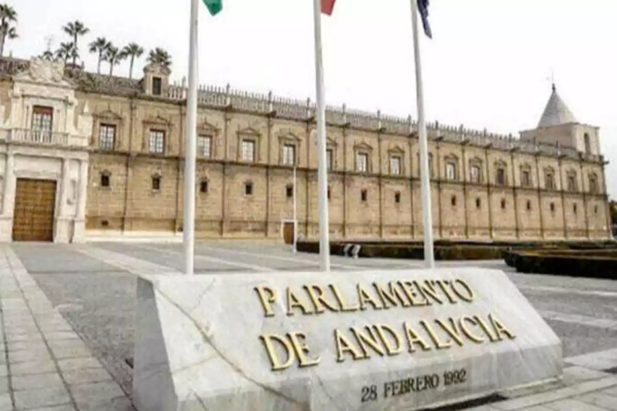 Edificio del Parlamento de Andalucía con una placa conmemorativa que dice "Parlamento de Andalucía 28 Febrero 1992" y tres banderas ondeando frente a él.
