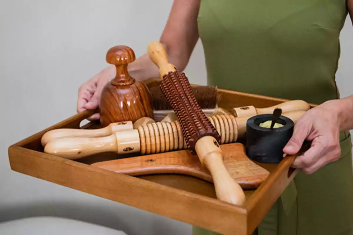 Persona sosteniendo una bandeja de madera con herramientas de masaje de madera y un recipiente pequeño.