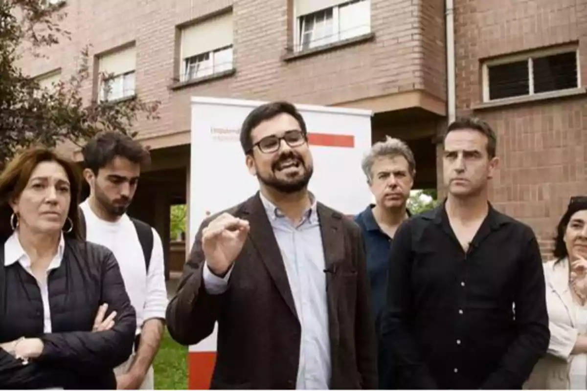 Un grupo de personas, algunas con expresión seria y otras hablando, se encuentra frente a un edificio de ladrillo.