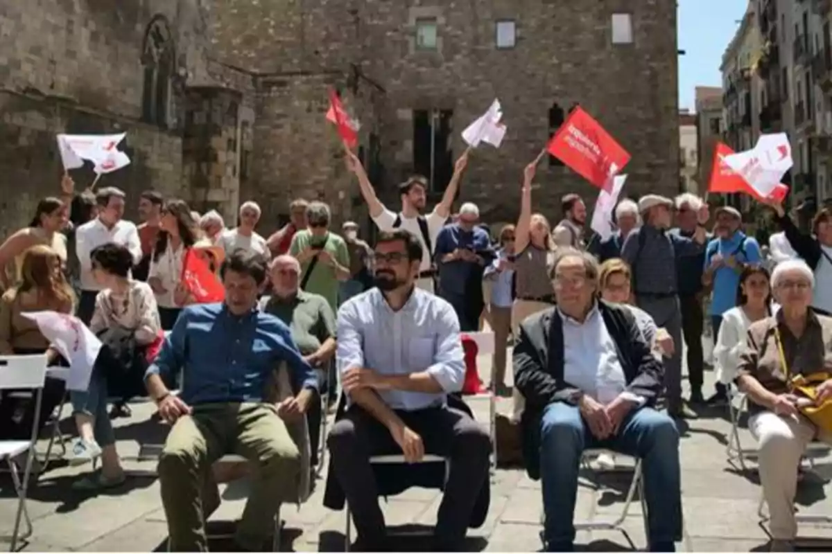 Un grupo de personas se encuentra reunido al aire libre, algunas sentadas y otras de pie, mientras varias de ellas sostienen y agitan banderas rojas y blancas.