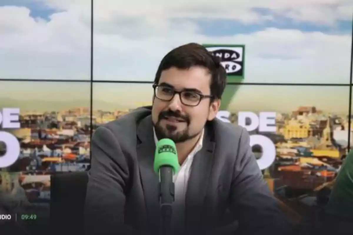 Un hombre con gafas y barba hablando en un programa de radio con un micrófono verde.