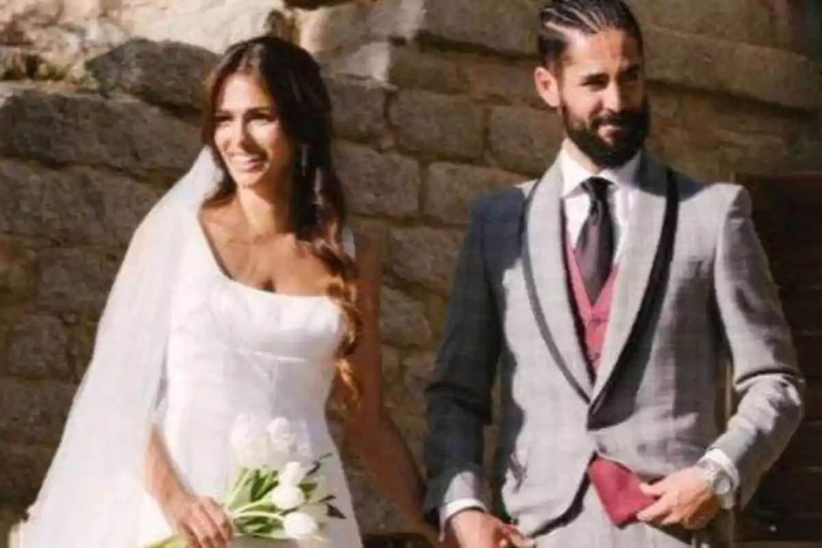 Una pareja sonriente vestida de novios, ella con un vestido blanco y un ramo de flores, y él con un traje gris y chaleco rojo, posan frente a una pared de piedra.
