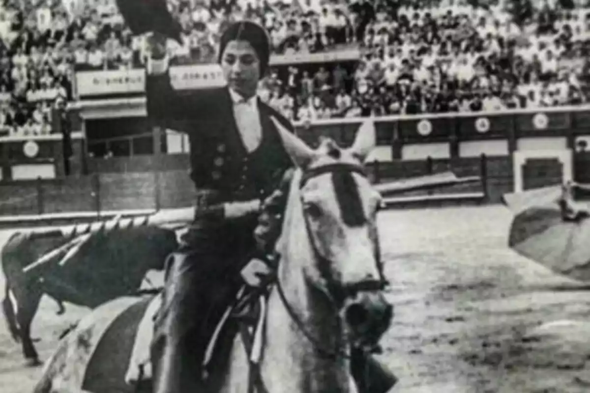 Una persona montada en un caballo en una plaza de toros, saludando al público con su sombrero en la mano.