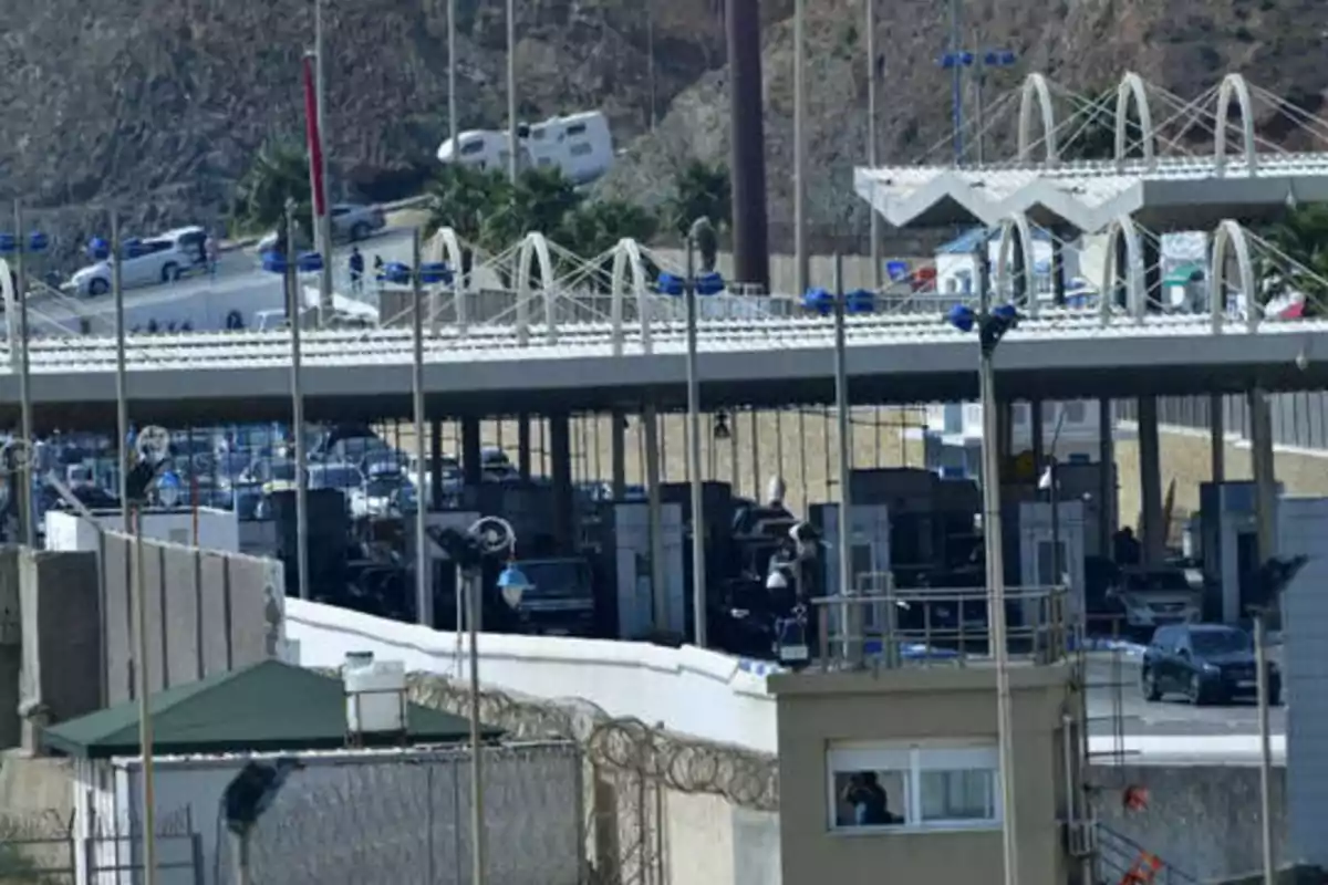 Imagen de un puesto fronterizo con varios carriles y vehículos pasando por controles de seguridad, con una estructura de techo y colinas en el fondo.