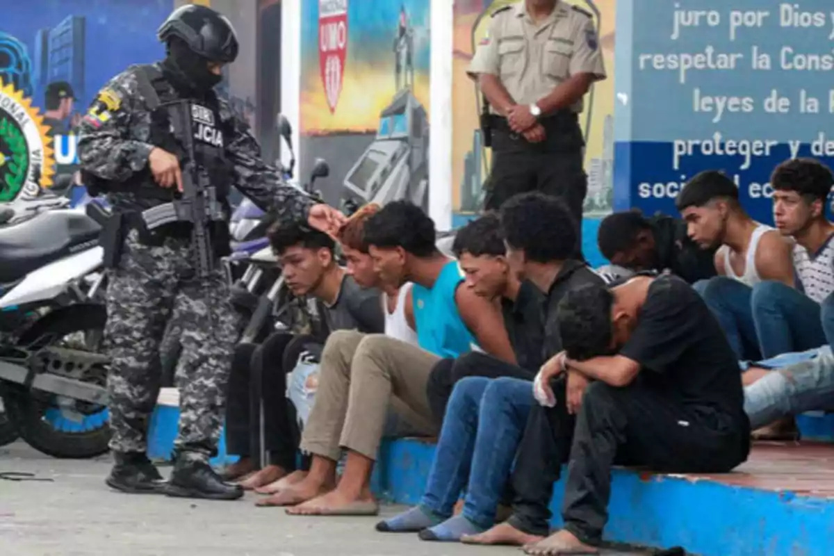 Un grupo de jóvenes sentados en el suelo, algunos con la cabeza baja, mientras un oficial de policía armado y con uniforme táctico los vigila y otro oficial está de pie al fondo.