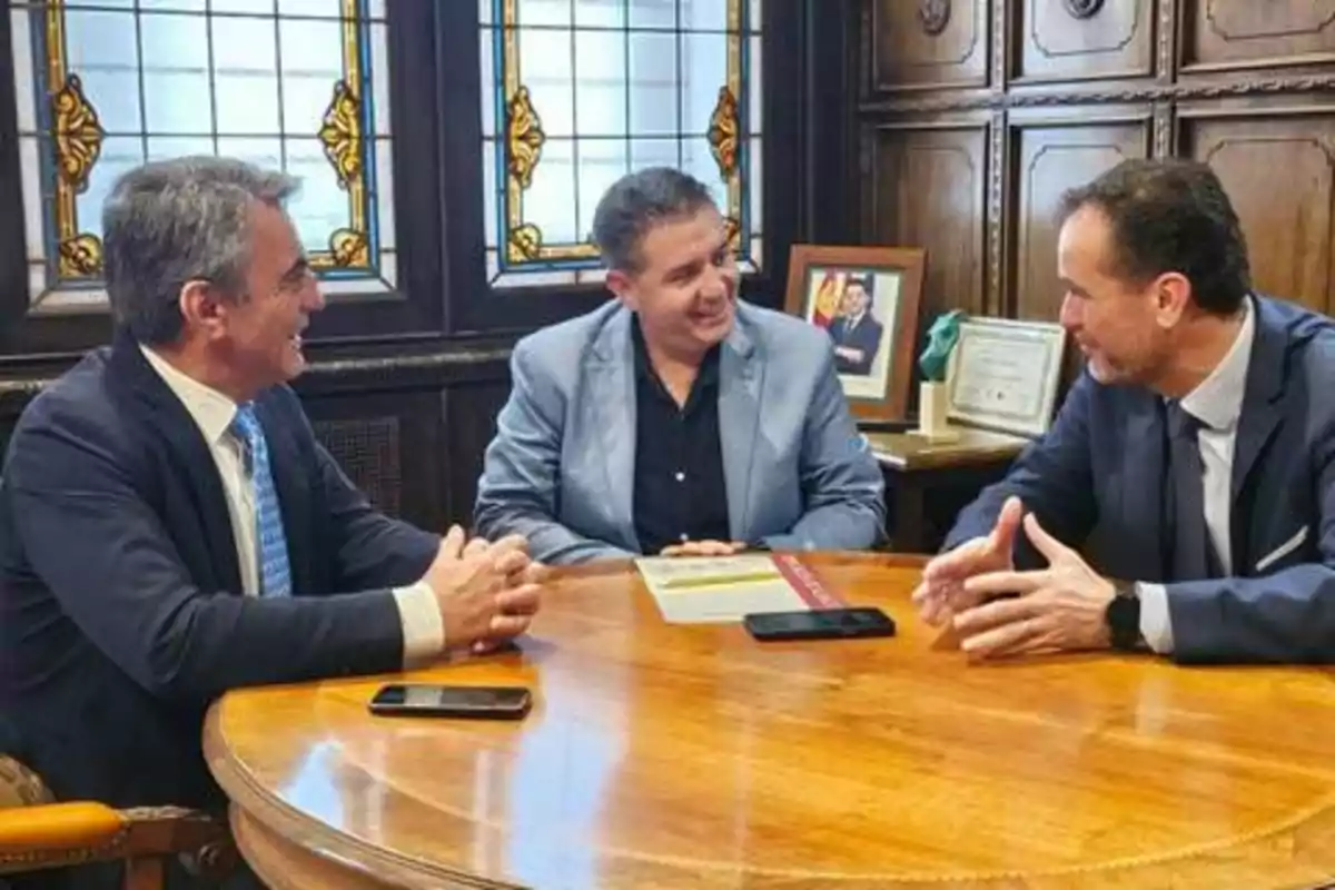 Tres hombres en trajes formales conversan alrededor de una mesa redonda de madera en una oficina con vitrales y muebles de madera.