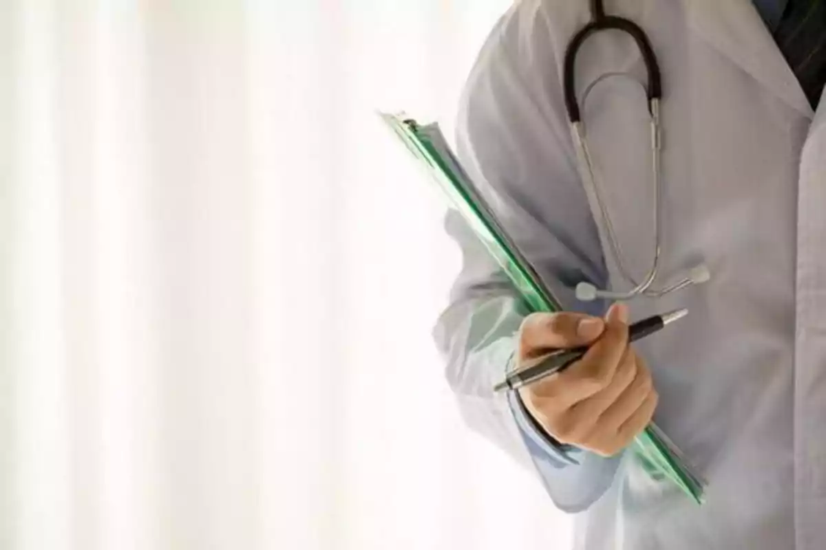 Un médico con bata blanca y estetoscopio sostiene una carpeta verde y un bolígrafo.