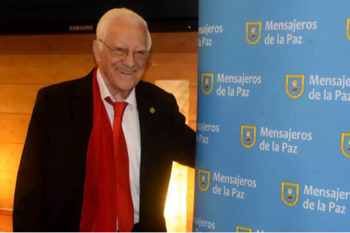 Un hombre mayor con gafas y corbata roja sonríe junto a un cartel azul que dice "Mensajeros de la Paz".