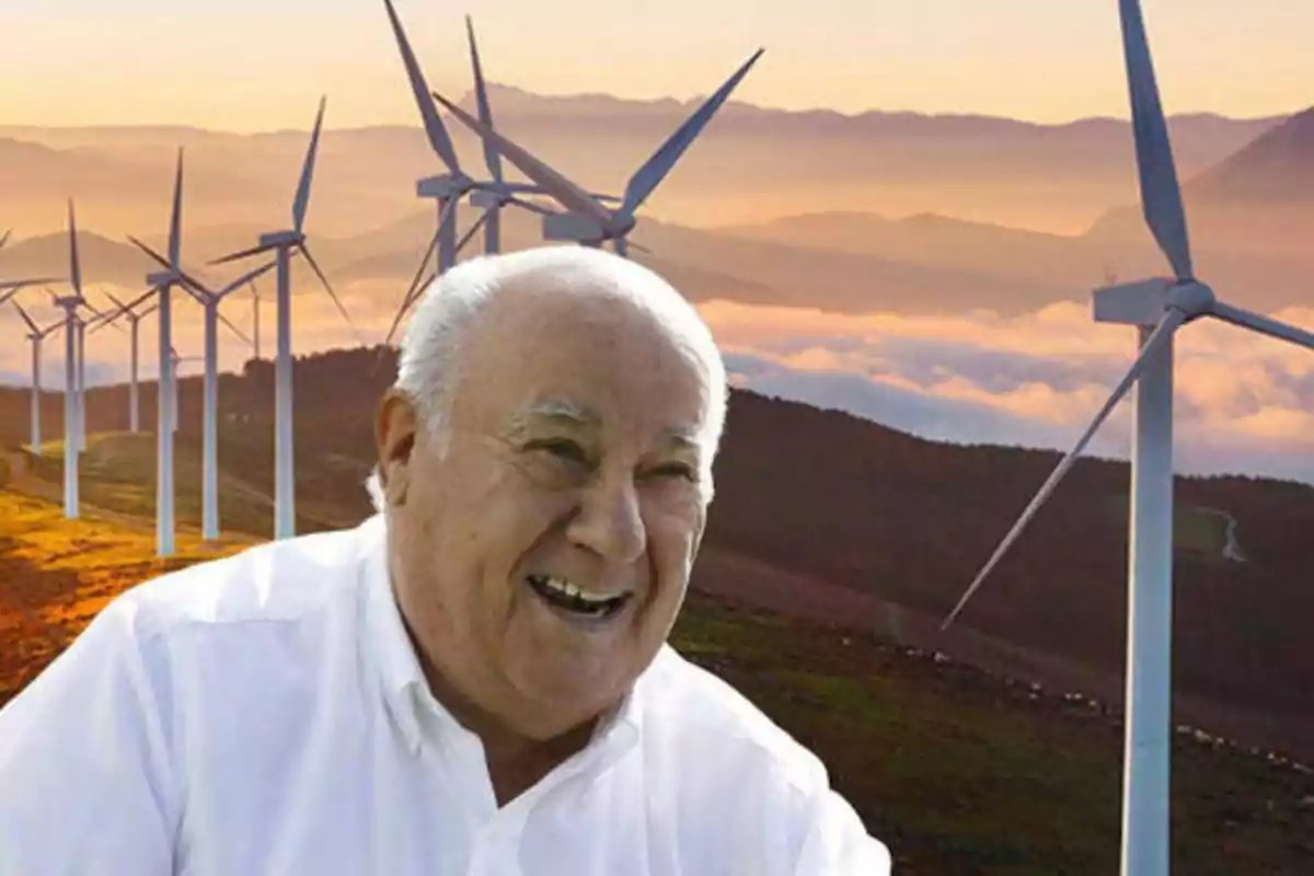 Un hombre mayor sonriente con una camisa blanca está en primer plano, mientras que en el fondo se ven varios aerogeneradores en un paisaje montañoso al atardecer.