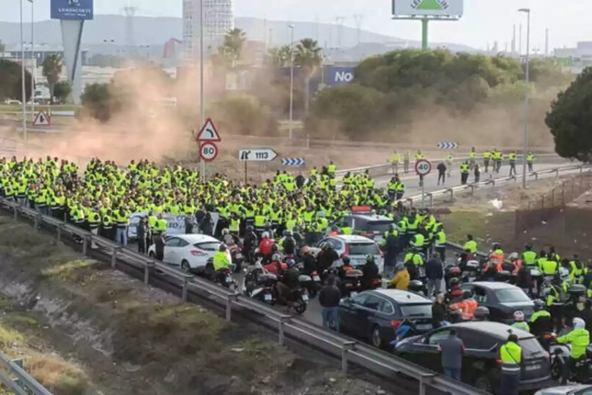 Una multitud de personas con chalecos amarillos bloquea una carretera mientras varios vehículos están detenidos.