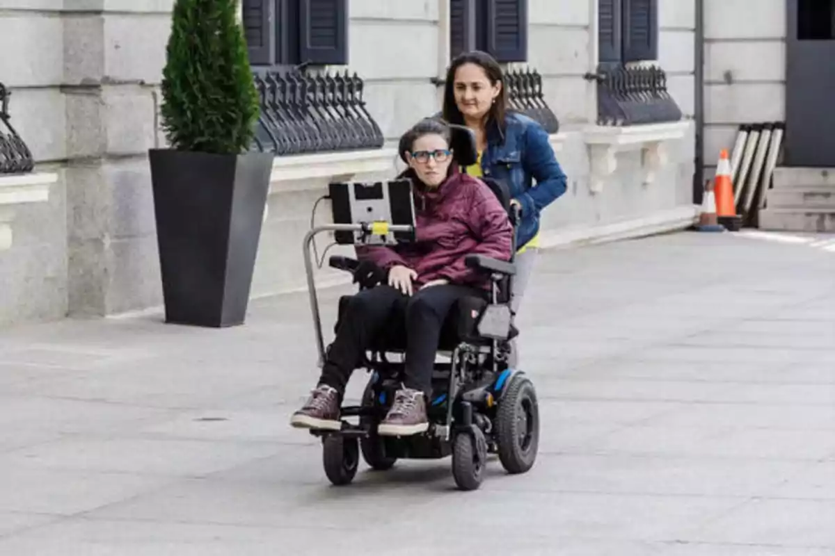 Una persona en silla de ruedas eléctrica siendo empujada por otra persona en una calle con edificios de fondo.