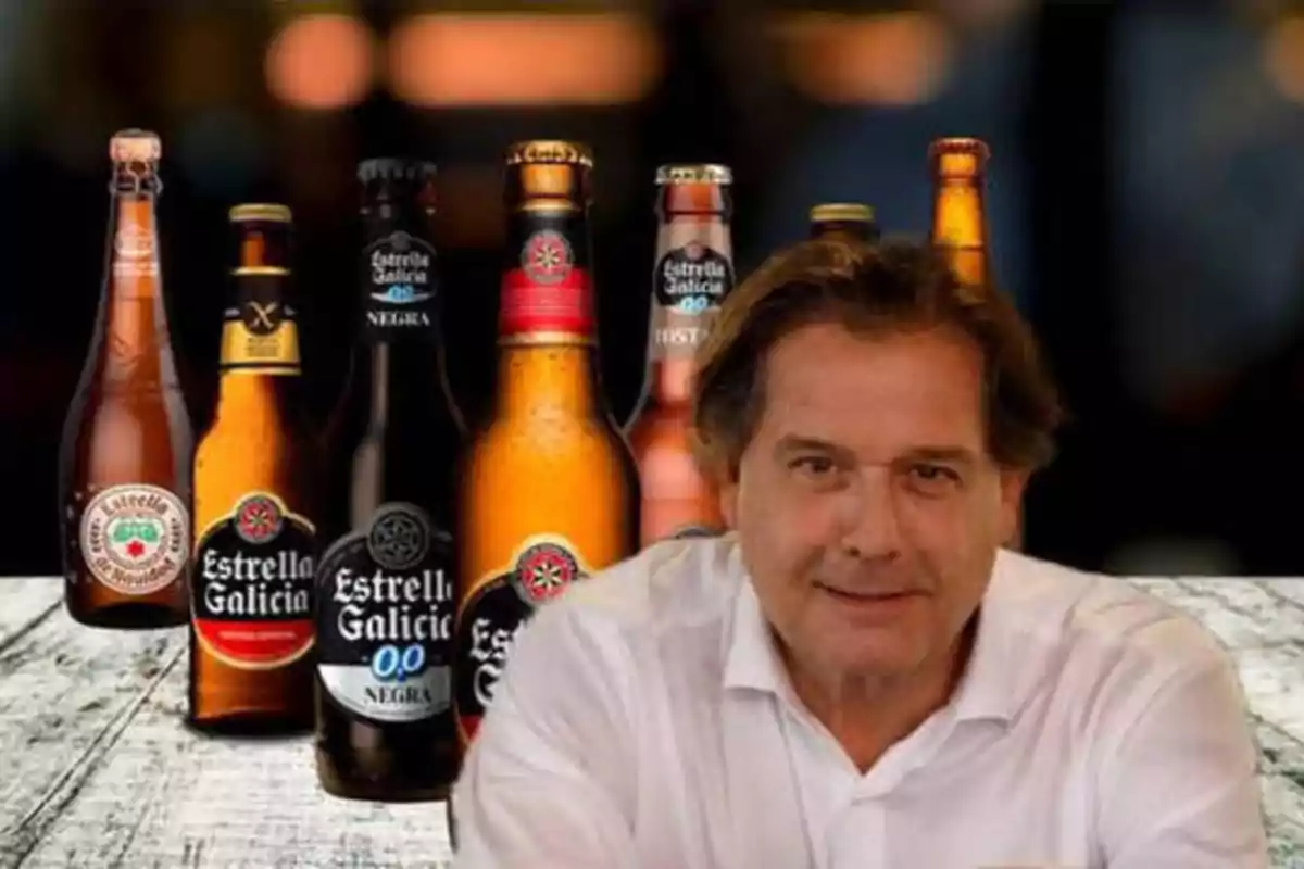 Un hombre con camisa blanca está sentado frente a una mesa de madera, detrás de él hay varias botellas de cerveza de la marca Estrella Galicia.