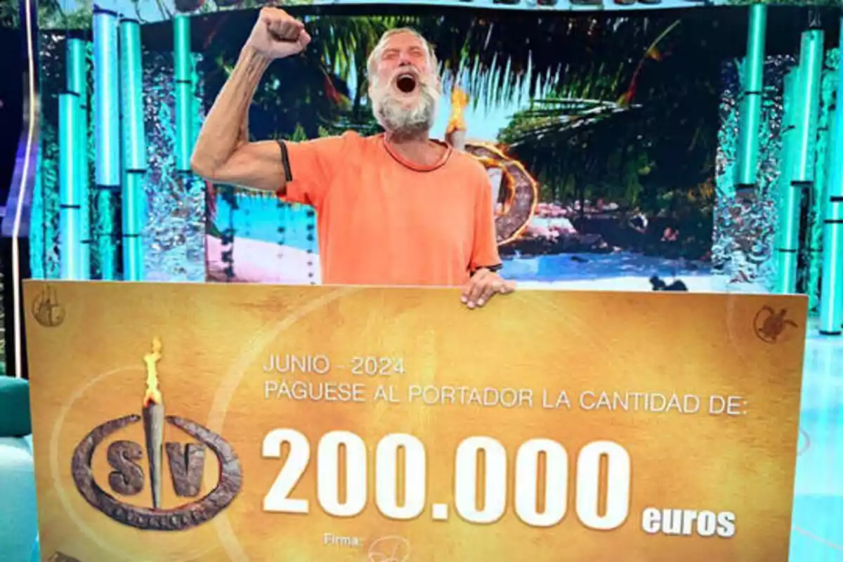 Un hombre con una camiseta naranja sostiene un cheque gigante por 200,000 euros, con una expresión de alegría y el puño levantado en señal de victoria.