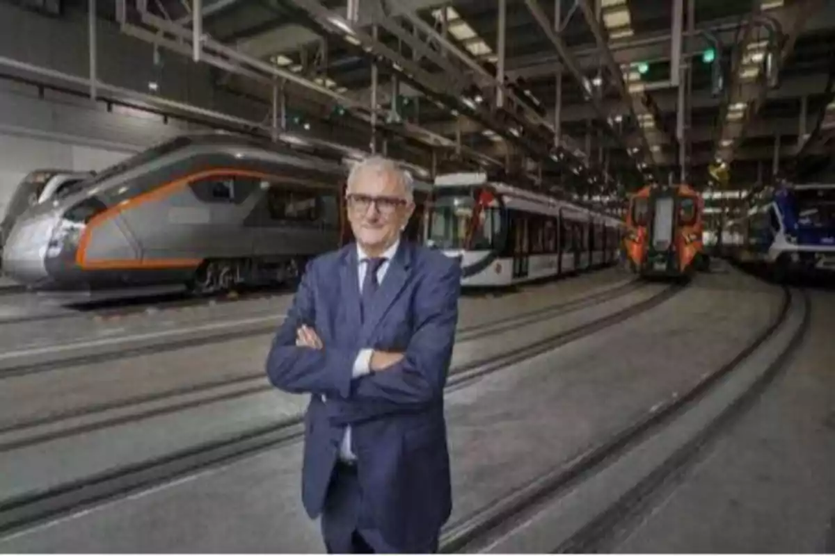 Un hombre de traje y gafas está de pie con los brazos cruzados en el interior de un taller de trenes, con varios trenes modernos en el fondo.