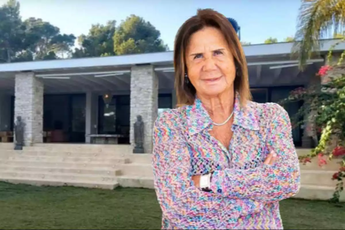 Una mujer de cabello castaño y camisa colorida está de pie con los brazos cruzados frente a una casa moderna con columnas de piedra y un jardín verde.