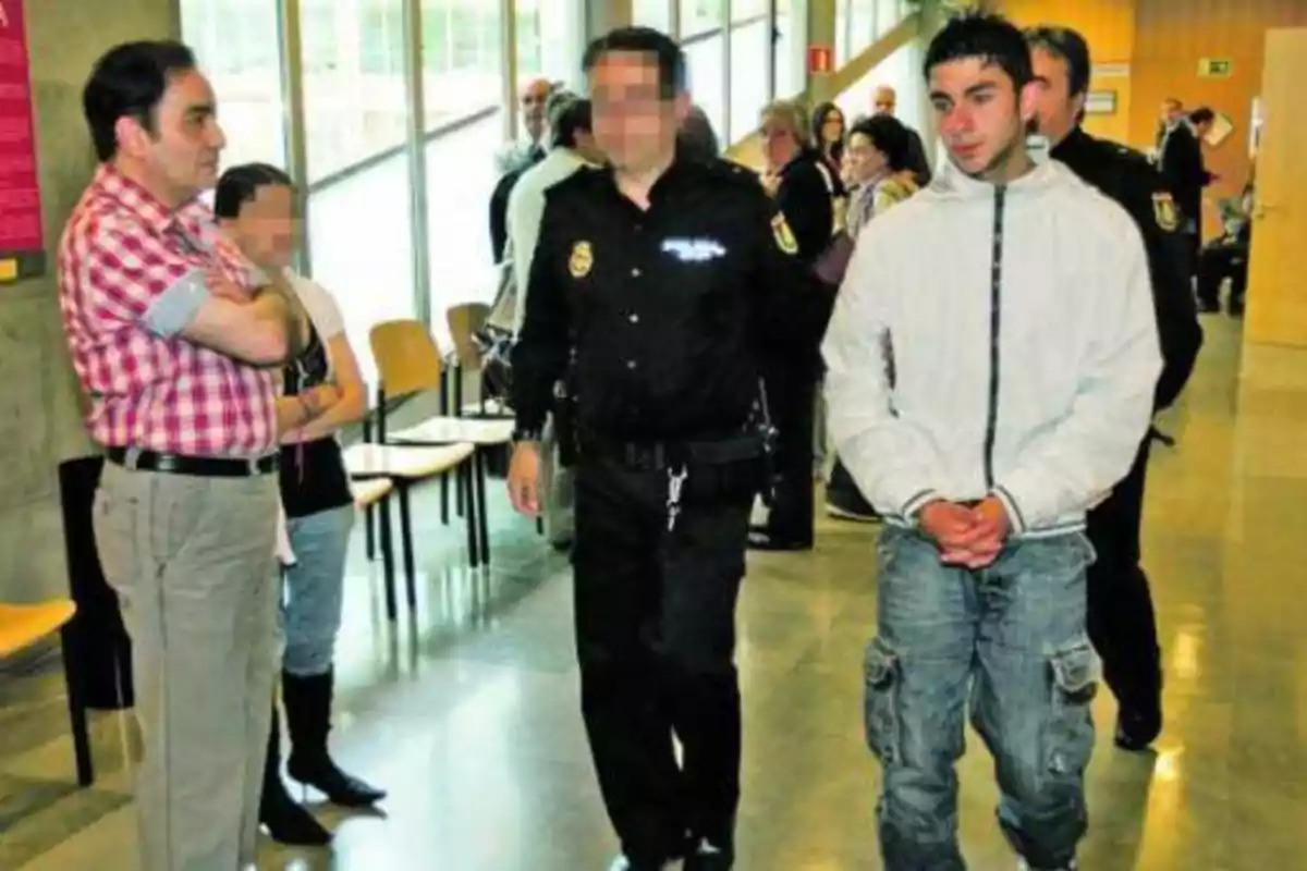 Un joven esposado es escoltado por un policía mientras otras personas observan en un pasillo.