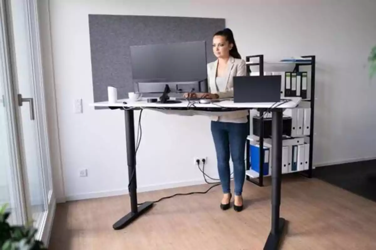 Mujer trabajando en una oficina con un escritorio de pie ajustable, usando una computadora y rodeada de estantes con carpetas y documentos.