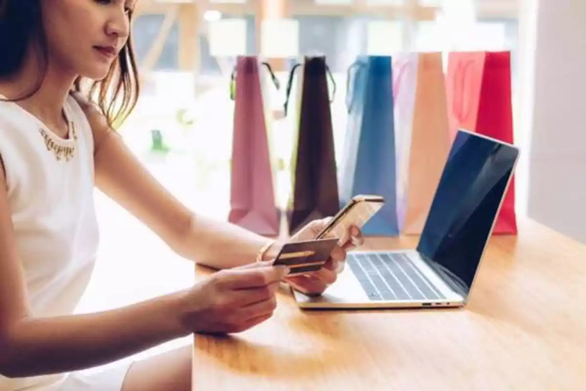 Mujer comprando en línea con una tarjeta de crédito y un teléfono móvil frente a una computadora portátil, con bolsas de compras en el fondo.